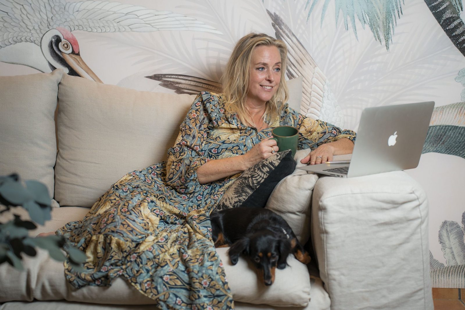 Sara Lee ler i en krämvit linnesoffa i en blå och gul blommig klänning, med en grön kopp i handen, laptopen framför sig och en tax som vilar bredvid henne.