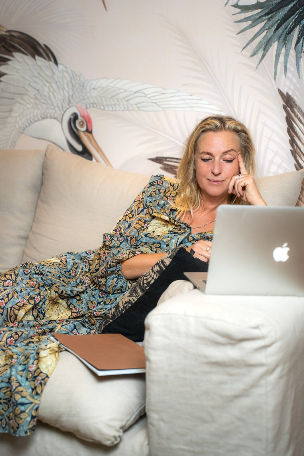 Sara Lee vilar i en krämvit linnesoffa i en blå och gul blommig klänning, med handen mot tinningen, och läser på sin laptop med ett milt leende. Bakom henne en målad trana och palmblad.